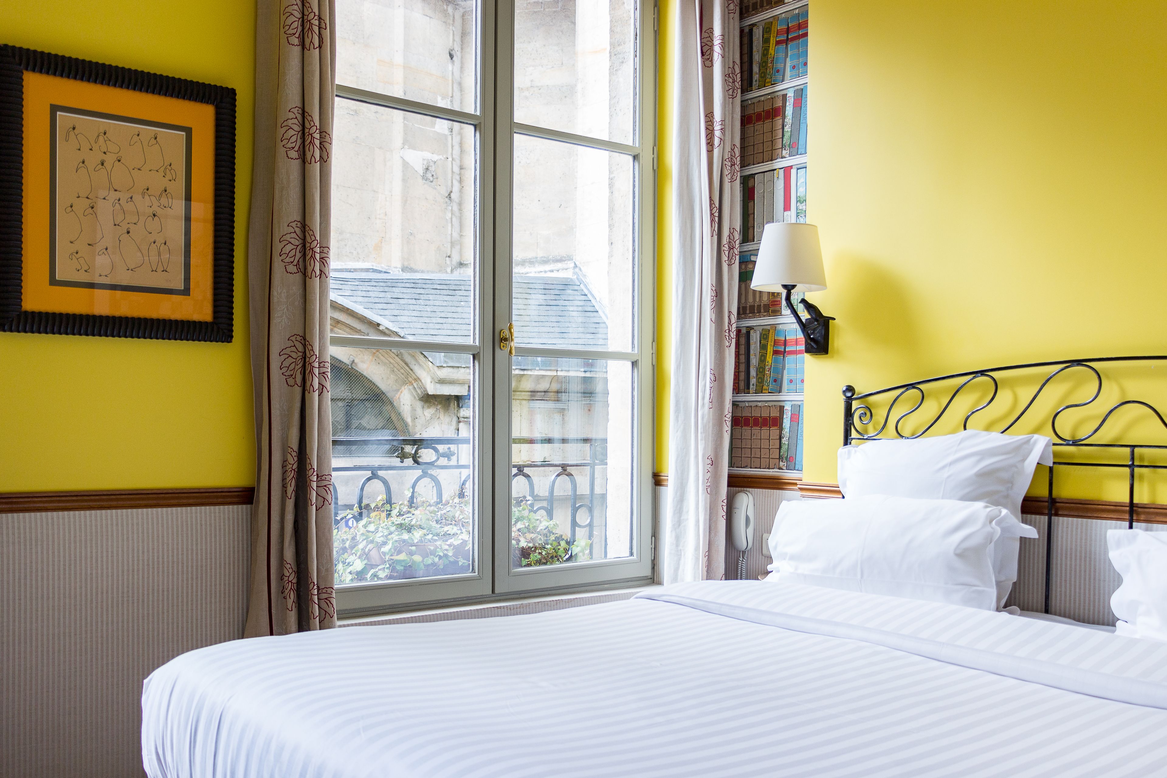 A hotel room with a large window, a bed with white linens, a bookshelf, and a framed picture on the wall.