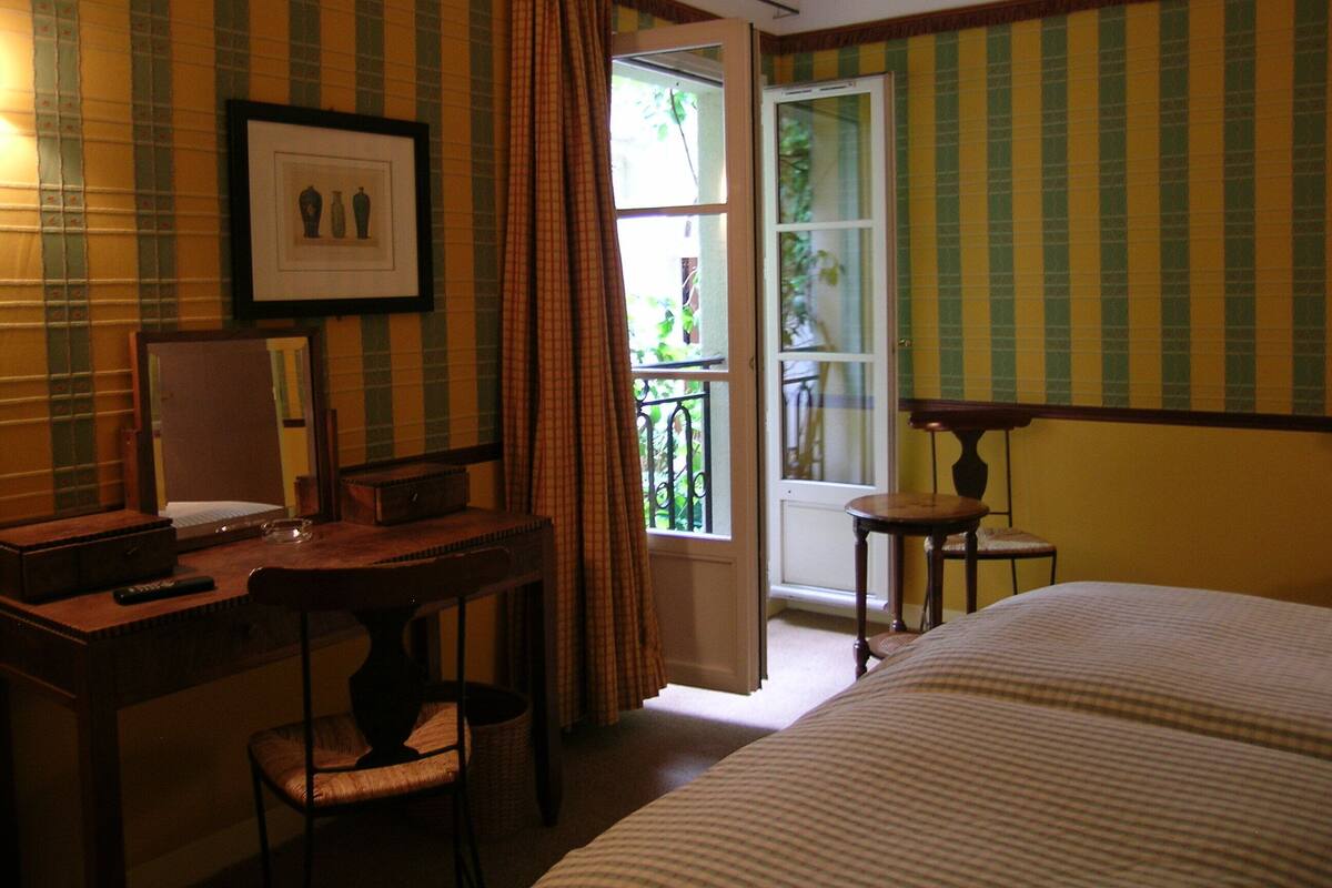 Una habitación de hotel con una cama, un escritorio con silla, un cuadro en la pared y una ventana con cortinas.