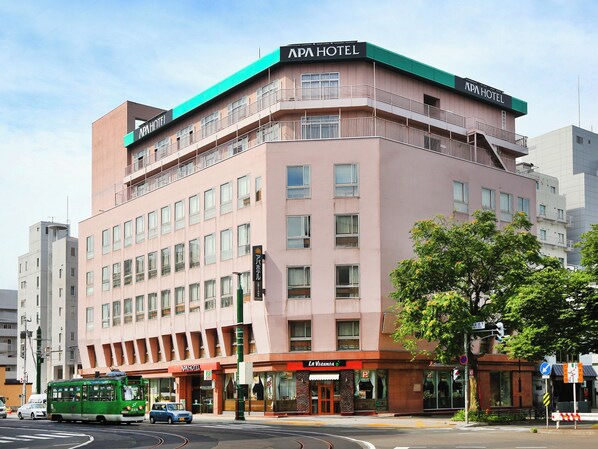 Um edifício com vários andares, com a placa APA HOTEL, um ônibus verde e uma faixa de pedestres.