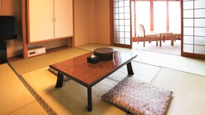 Une pièce de style japonais traditionnel, avec un sol recouvert de tatamis, une petite table basse avec un coussin, des portes coulissantes et un t éléviseur posé sur une étagère.