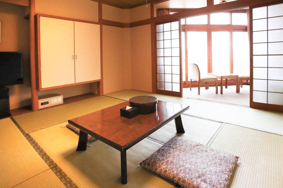 A traditional Japanese room with tatami flooring, a low table with a cushion, sliding doors, and a television on a shelf.