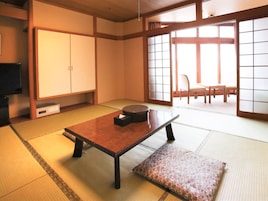 Una habitación japonesa tradicional con suelo de tatami, una mesa baja con cojín, puertas correderas y un televisor sobre una estantería.