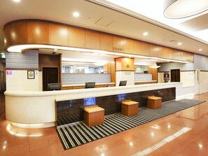 Uma área de recepção de hotel com balcão curvo, mesa de recepção e sala de espera com banquetas.