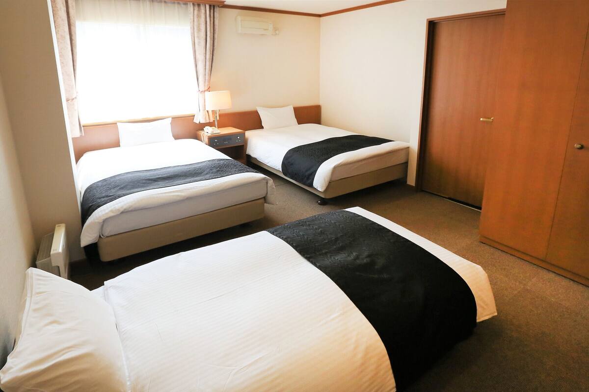 A hotel room with two beds, a bedside table with a lamp, a wardrobe, and a window with curtains.