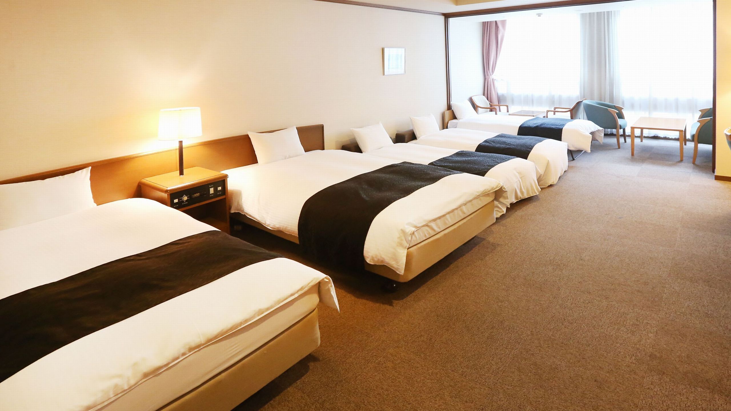 A hotel room with four beds arranged in a row, a desk with chairs, and a large window with curtains.