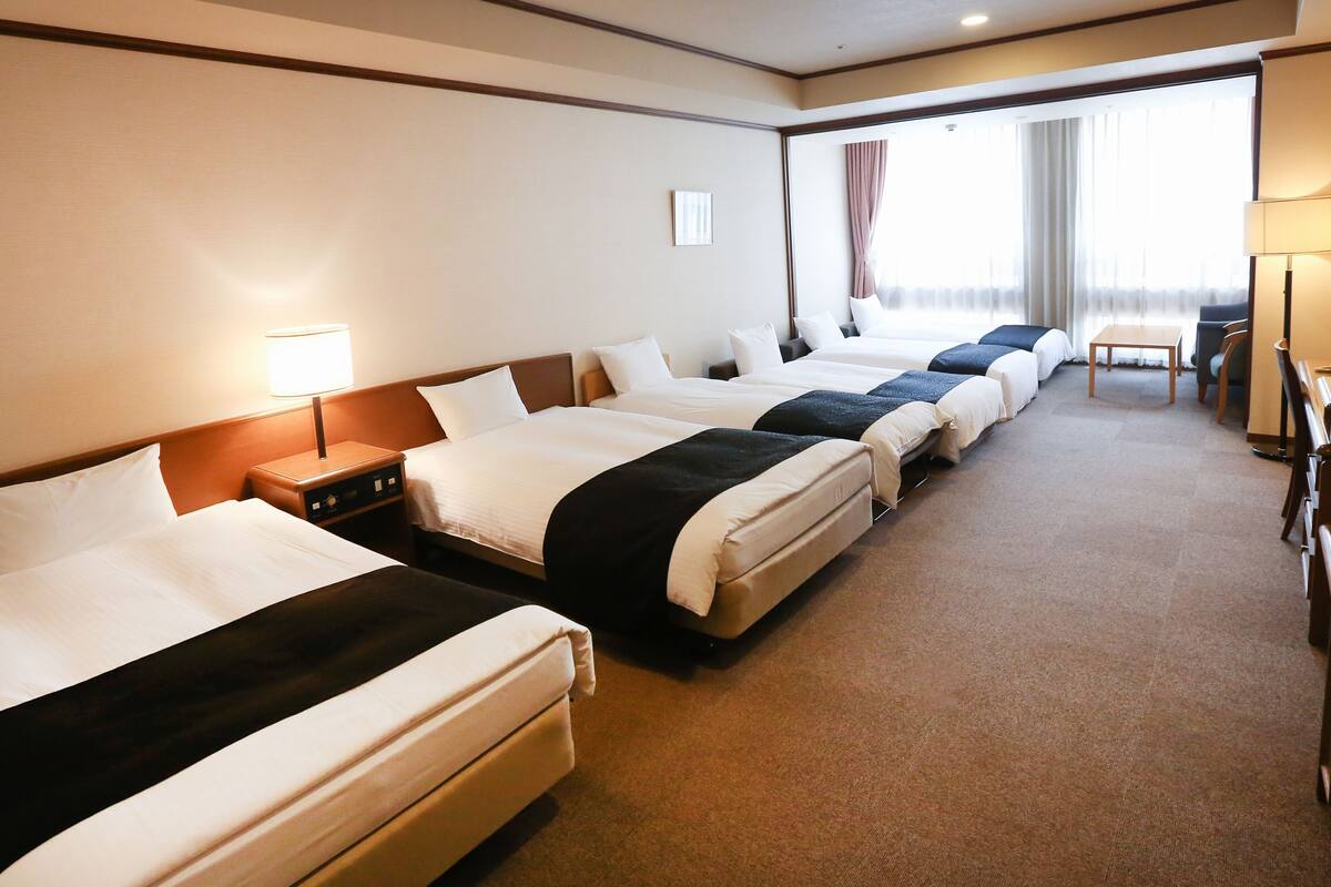 A hotel room with four beds in a row, a desk with a chair, and a window with curtains.