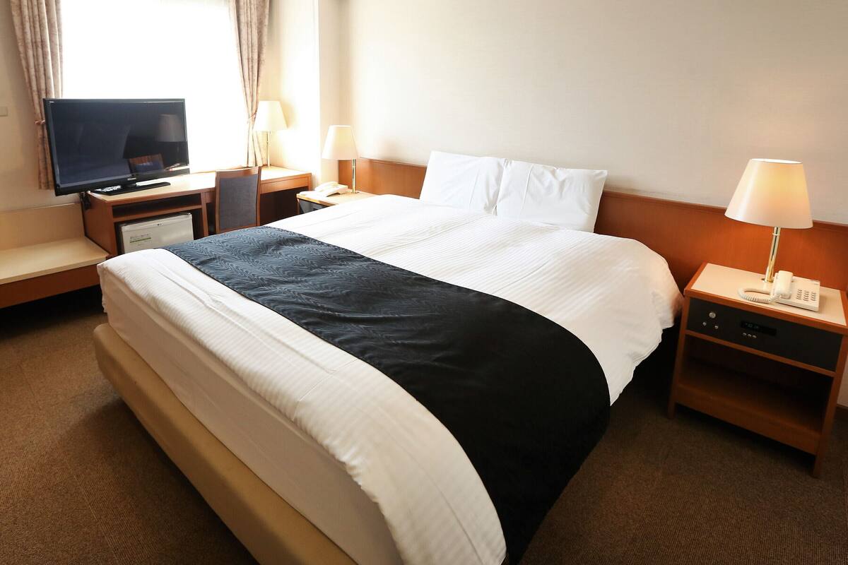 A hotel room with a bed, a desk with a television, two bedside lamps, a nightstand with a phone, and a window with curtains.