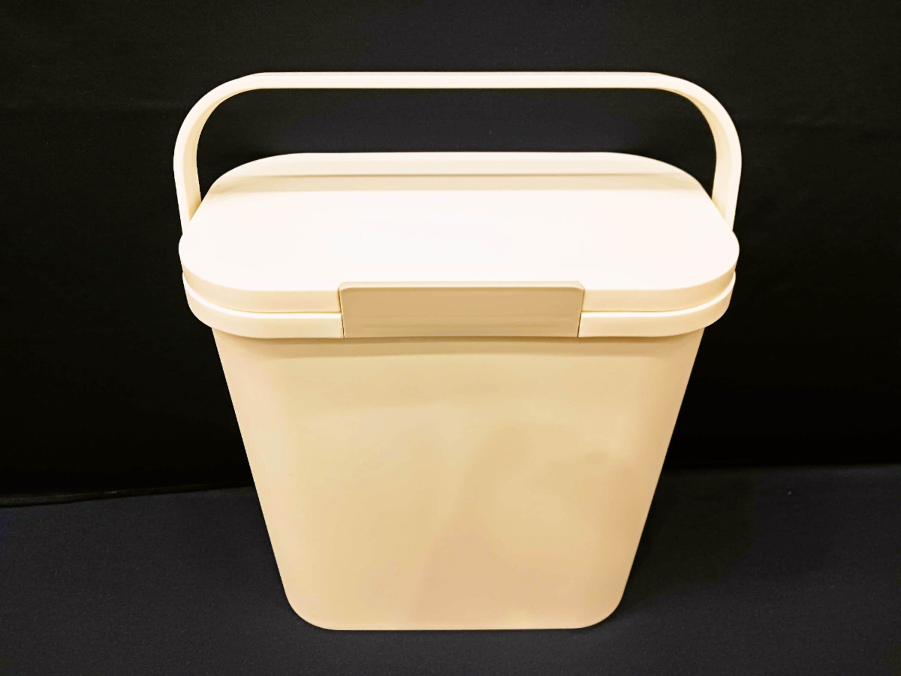 Un seau en plastique beige, doté d’une anse et d’un couvercle, posé sur une surface noire.
