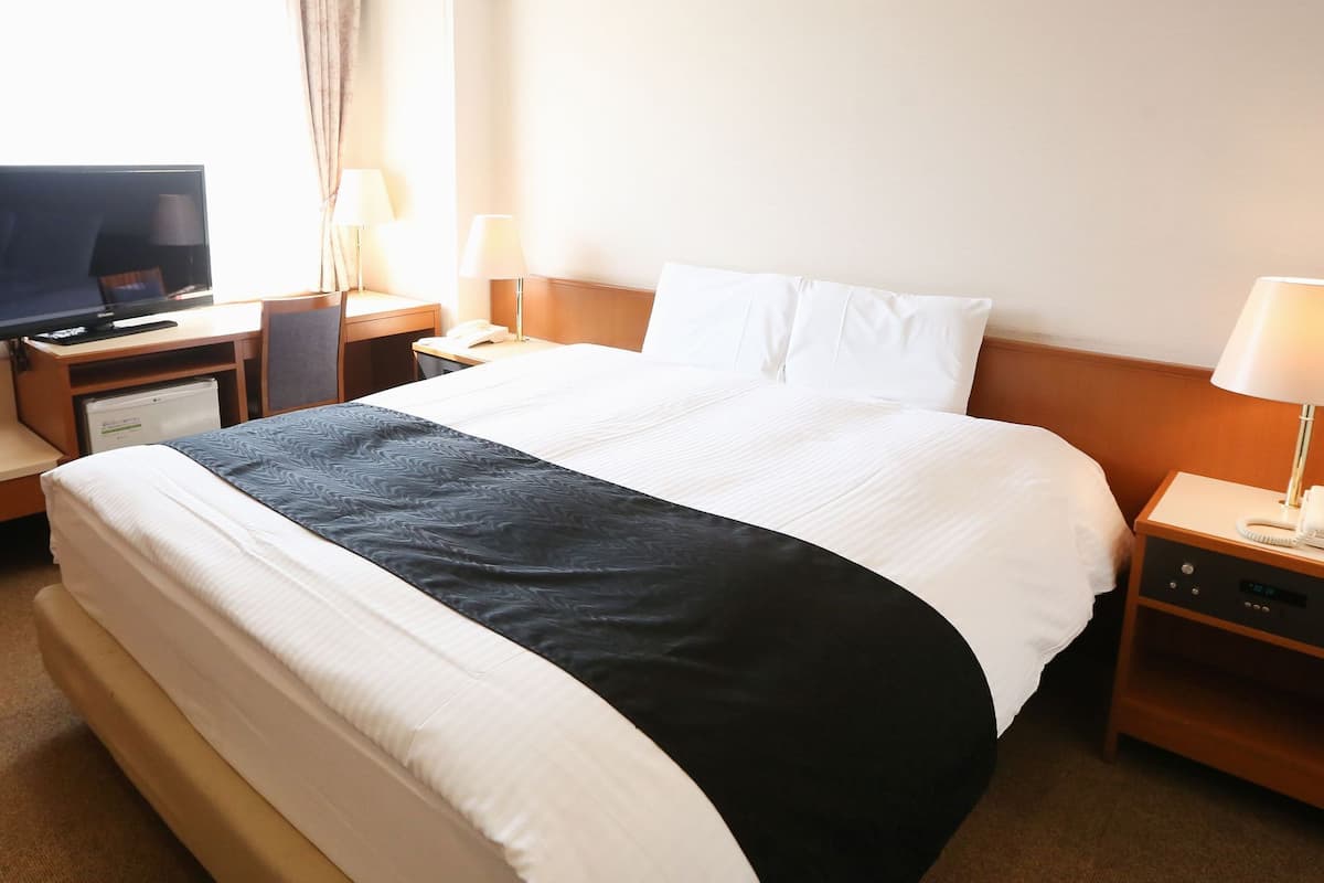 A hotel room with a bed, a desk with a television, a lamp, a phone, and a small refrigerator.