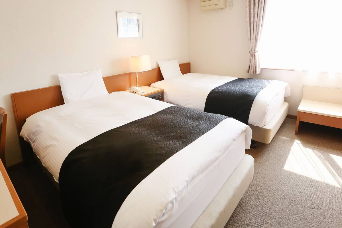 A hotel room with two beds, a bedside table with a lamp, an air conditioning unit, and a window with curtains.
