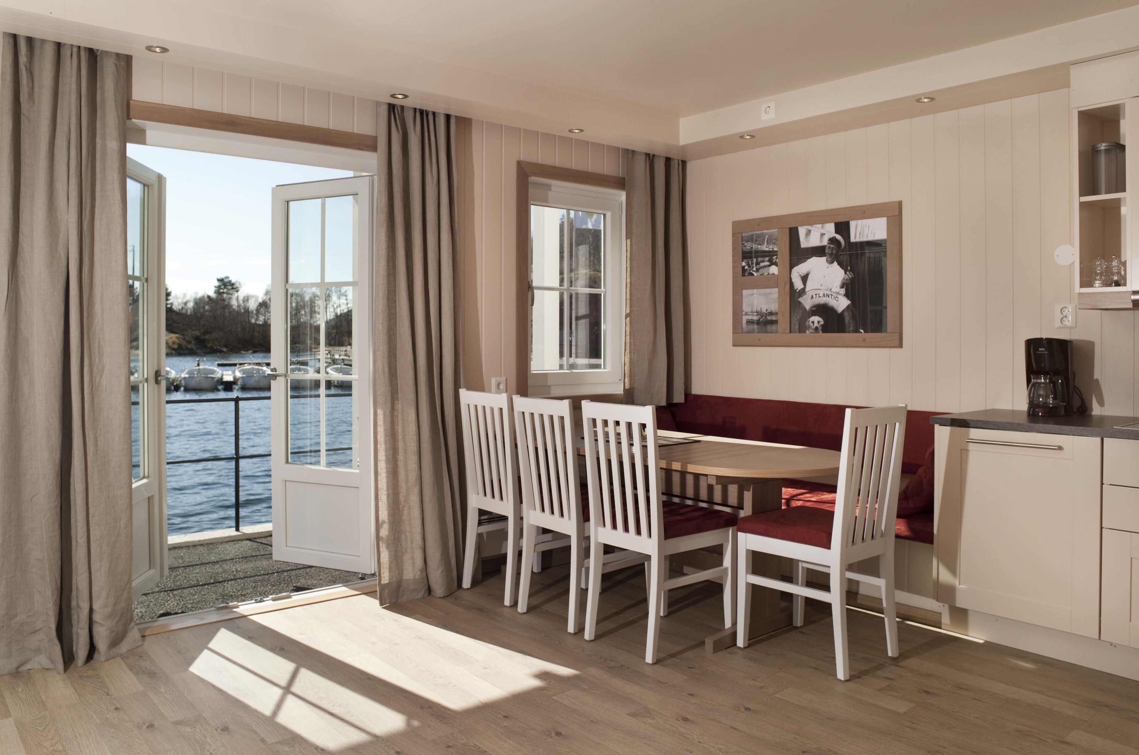 Un coin repas avec une table en bois et des chaises blanches, un banc rouge avec coussin, et une vue sur l’eau grâce à de grandes fenêtres.
