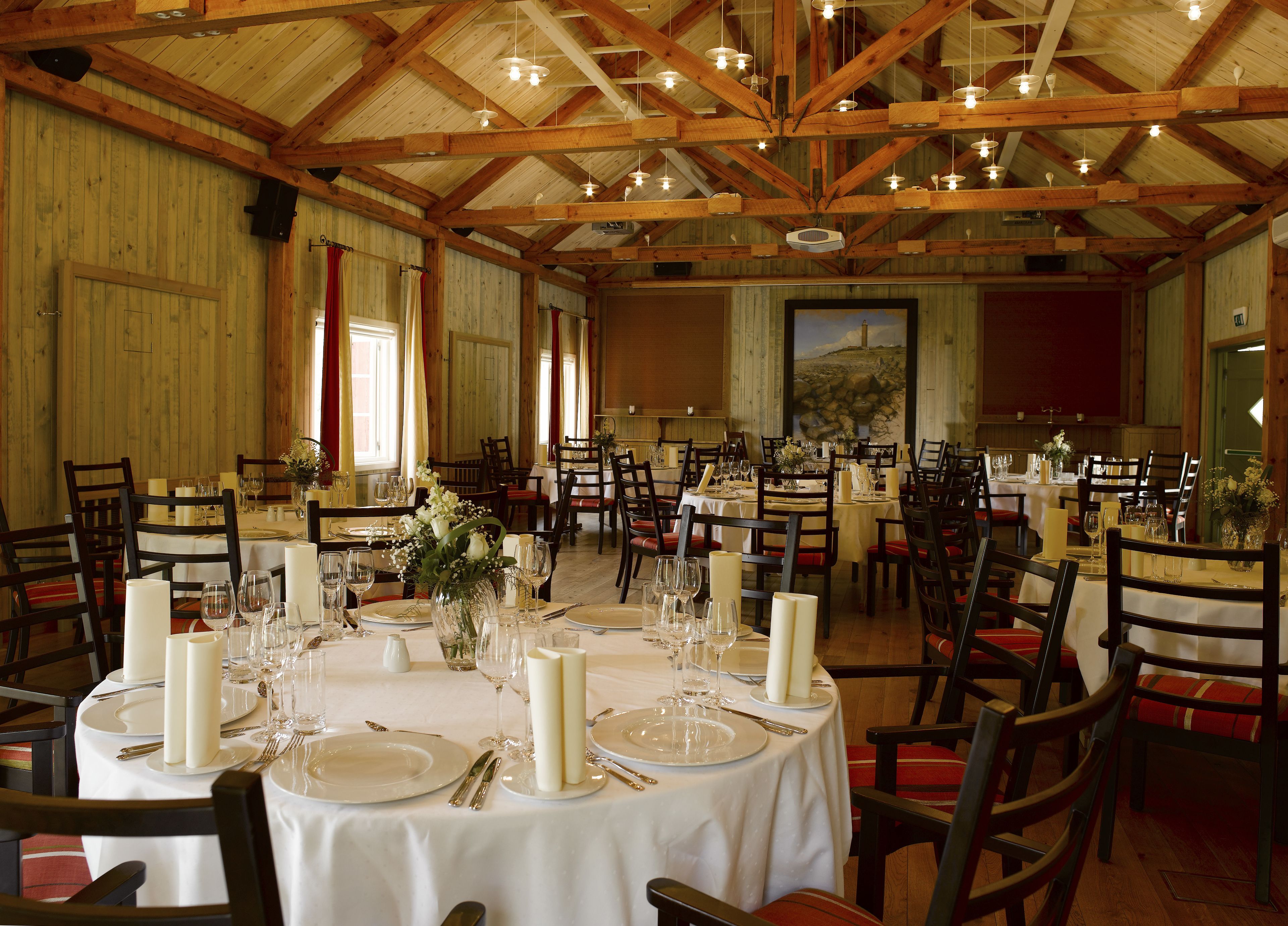 A large dining hall with multiple round tables set for a formal meal, wooden beams on the ceiling, and a large painting on the wall.