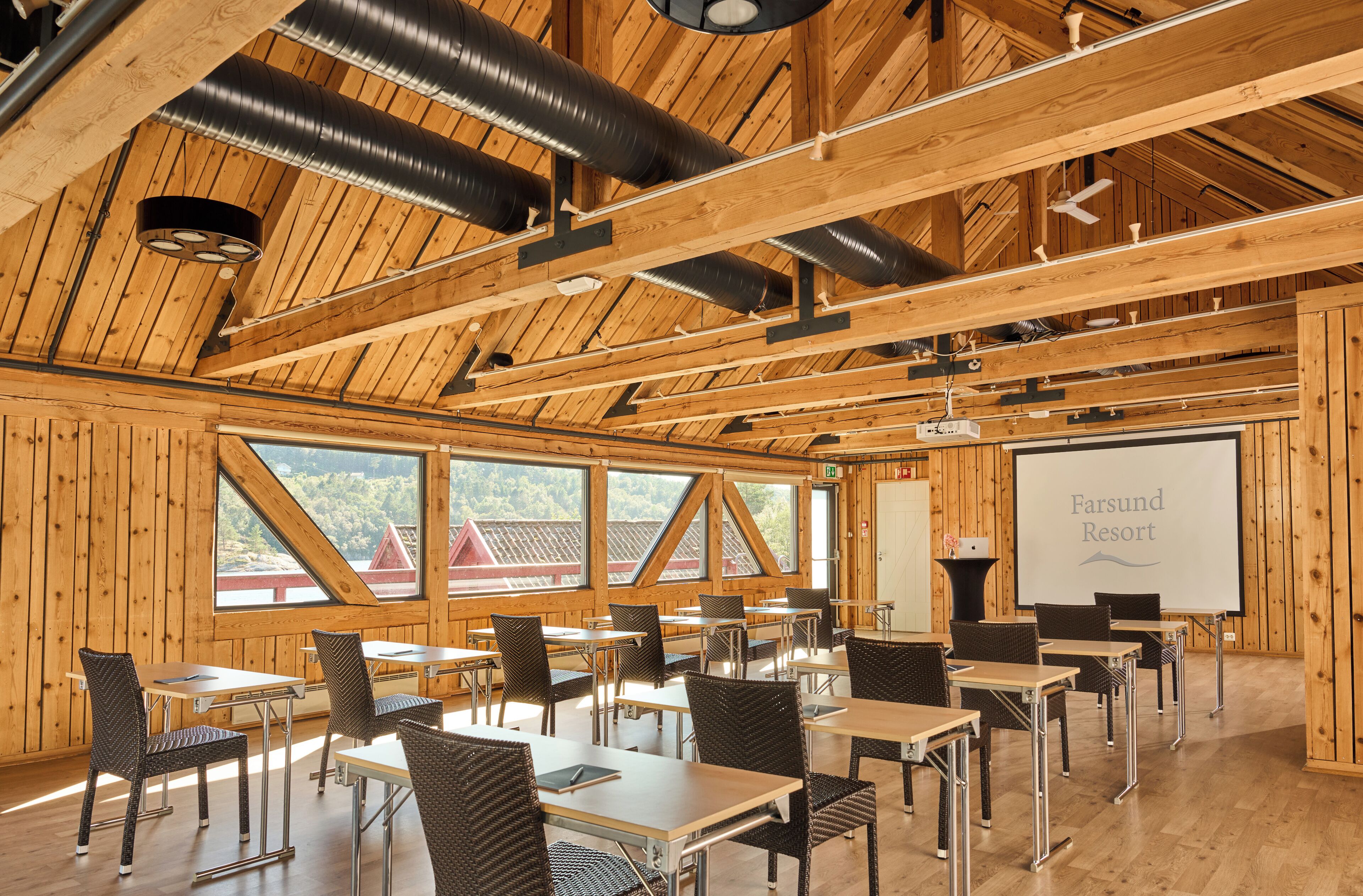 A conference room with tables and chairs, a projector screen displaying 'Farsund Resort', and large windows with a view of a red-roofed building.