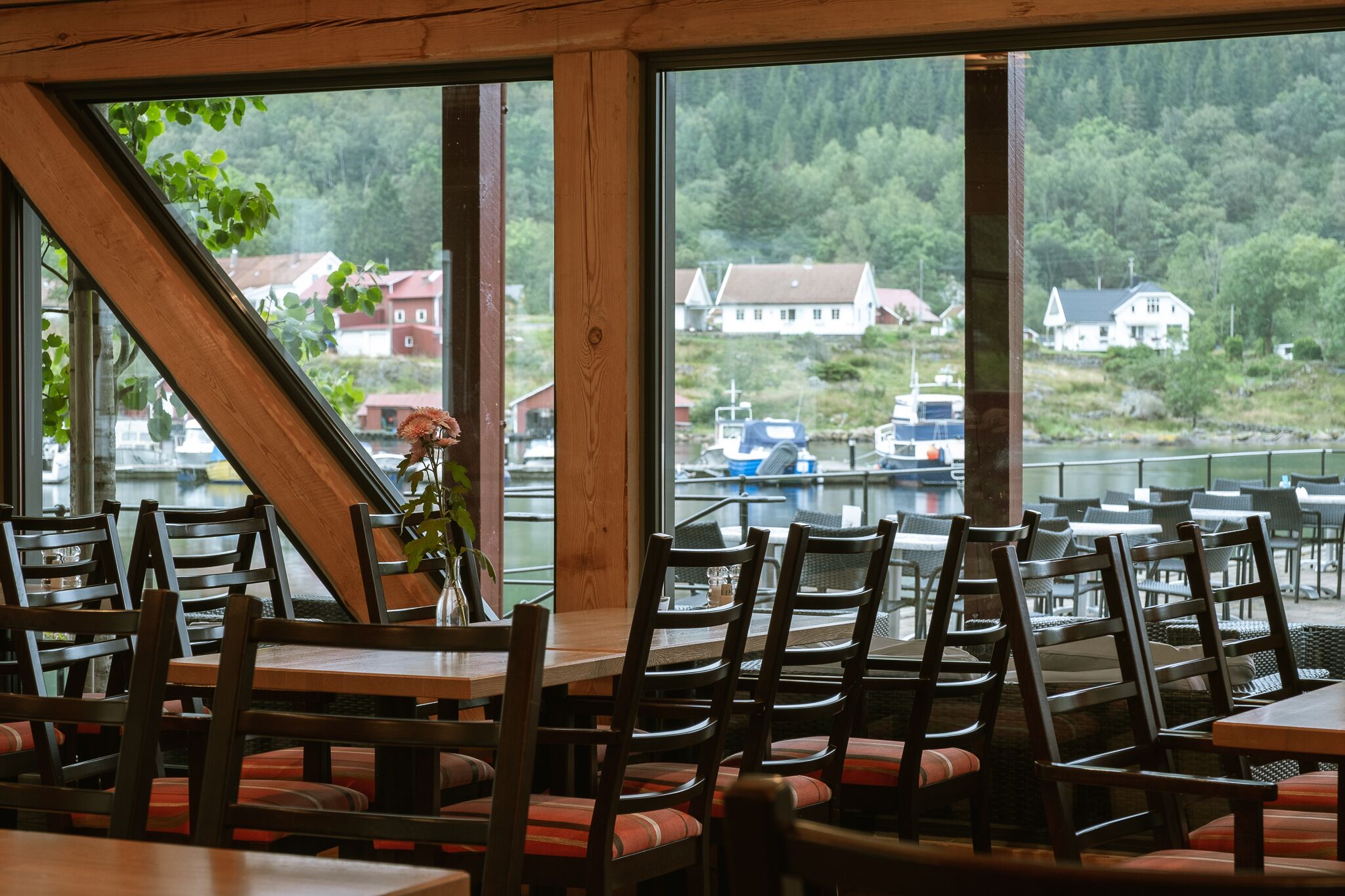 Un restaurante con mesas y sillas de madera, amplios ventanales con vista a un lago y casas.