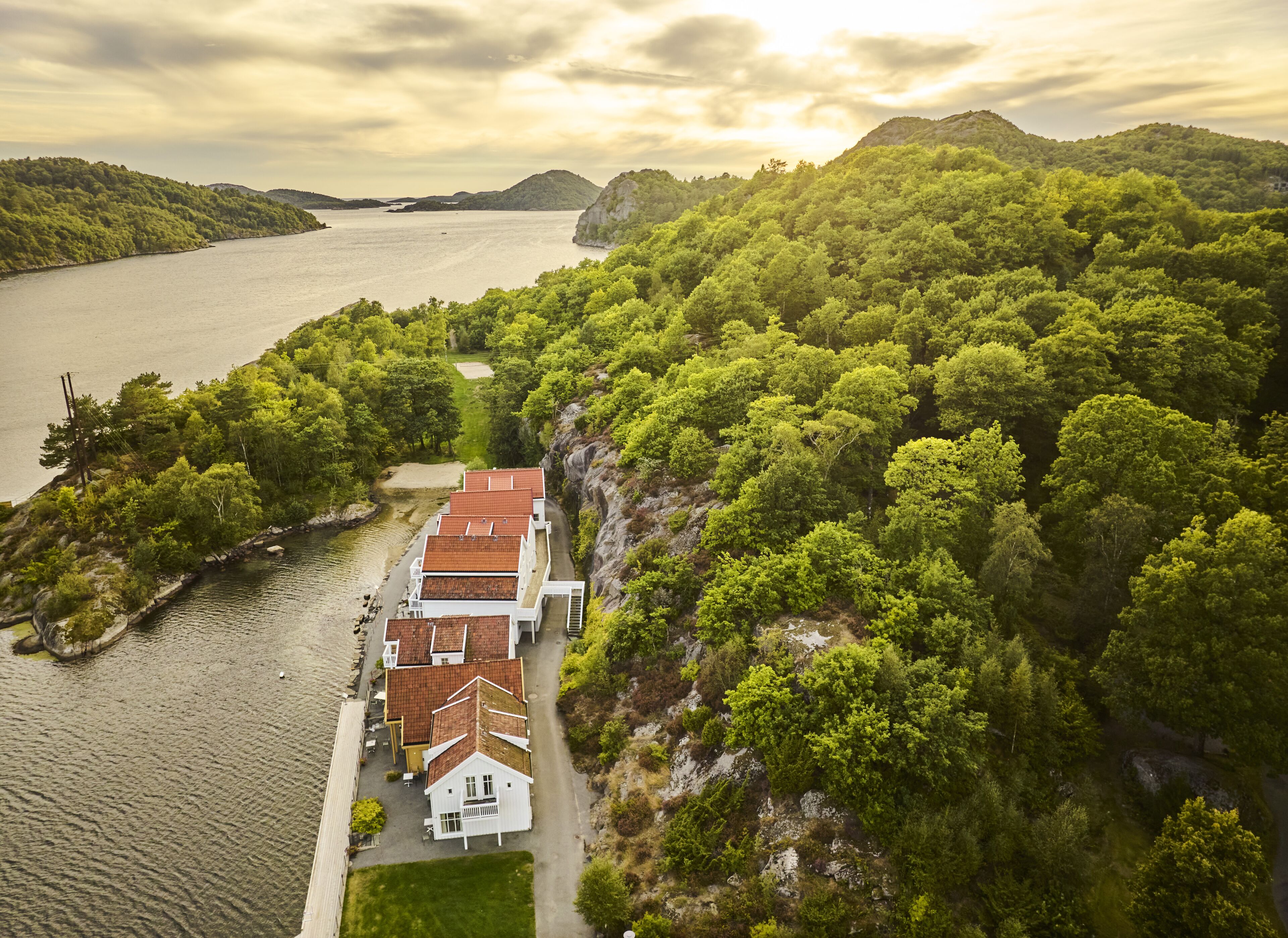 A coastal property with a white building featuring a red roof, surrounded by lush greenery and a body of water.