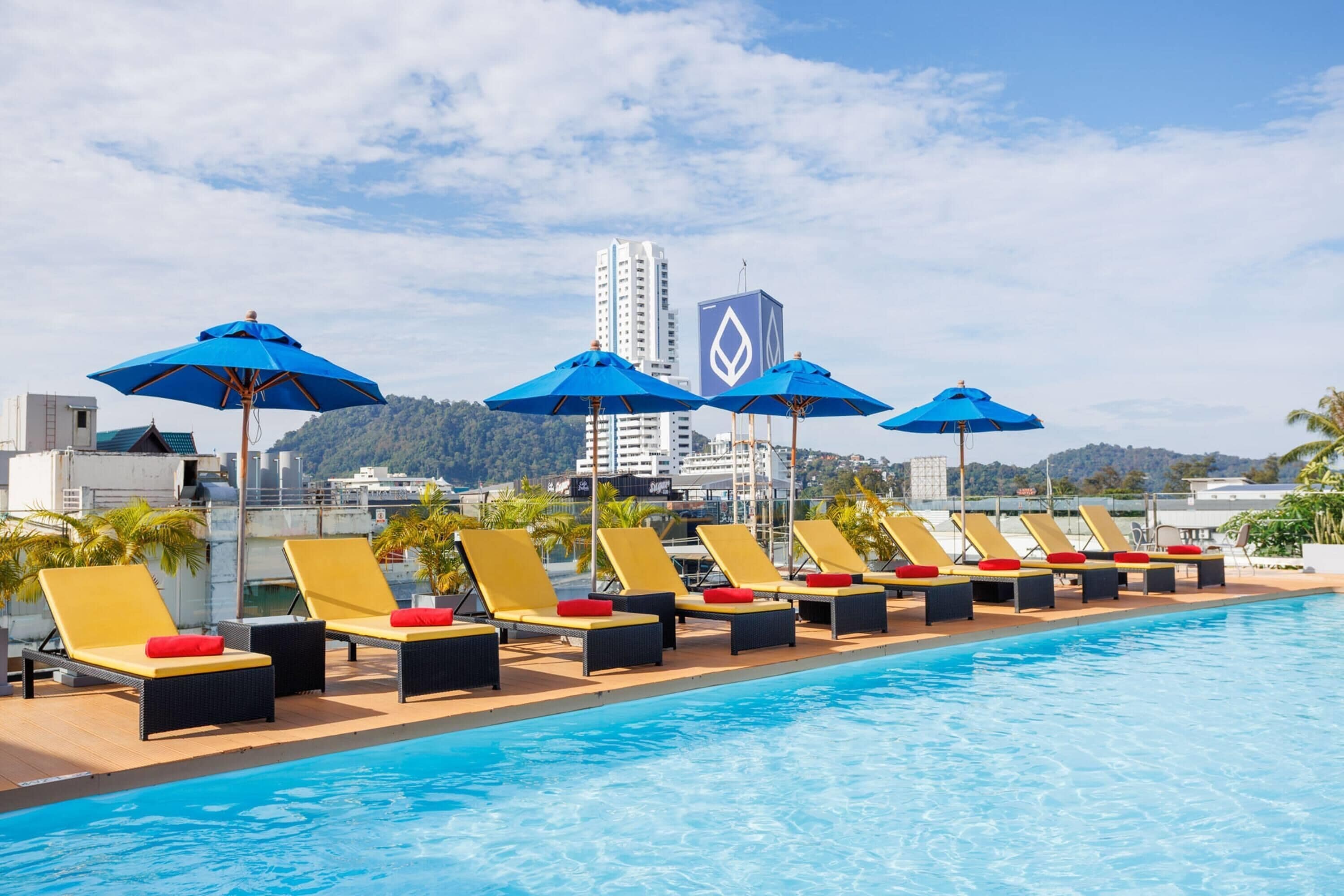 Zona junto a la piscina con tumbonas y sombrillas, cielo despejado y vista panorámica de un paisaje urbano con un edificio alto.