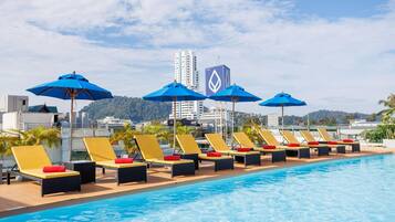 Zona junto a la piscina con tumbonas y sombrillas, cielo despejado y vista panorámica de un paisaje urbano con un edificio alto.