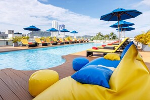 Un espace aménagé au bord de la piscine, avec des chaises longues et des parasols, une terrasse en bois et une vue sur les bâtiments en arrière-plan.