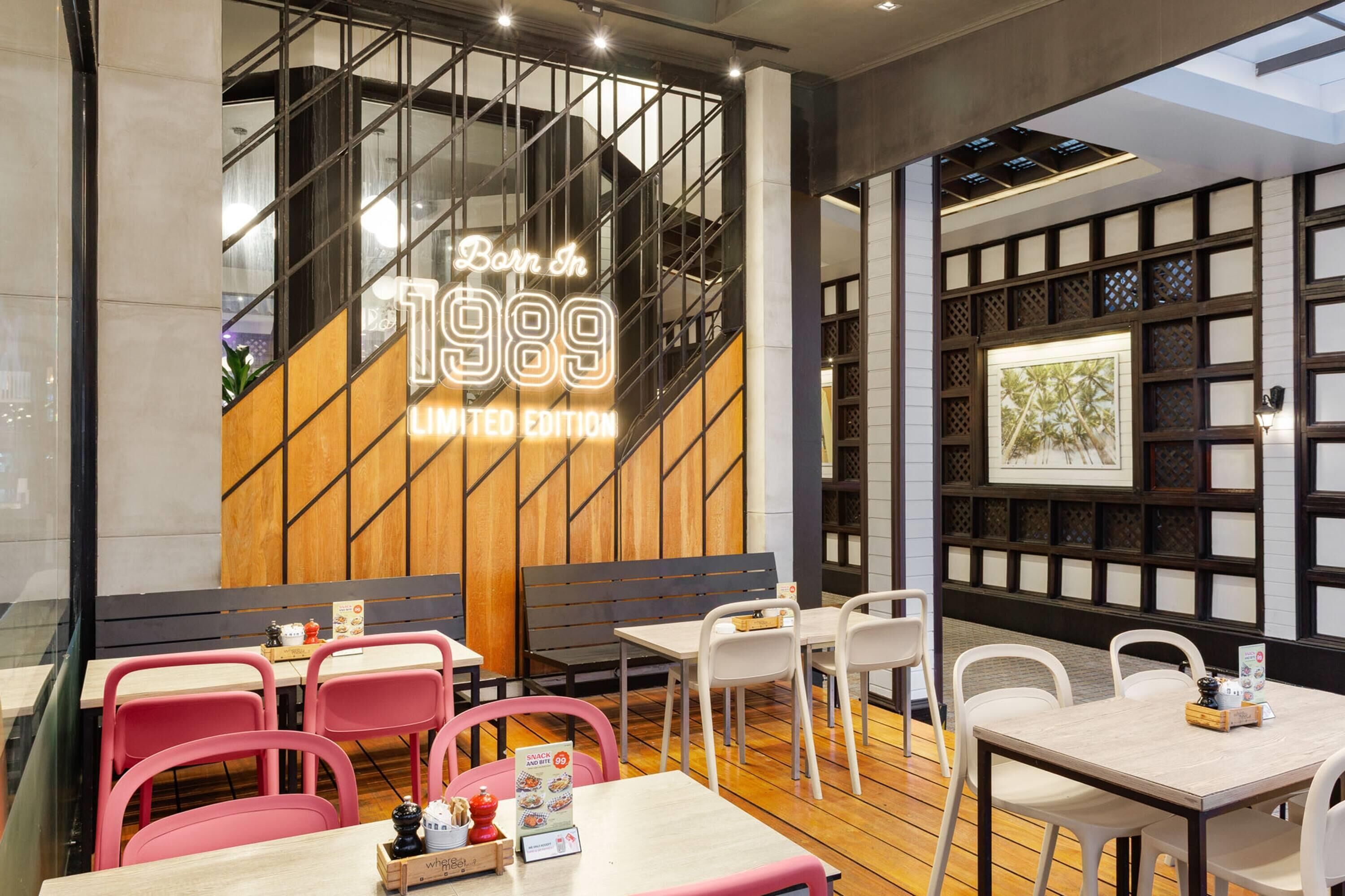 A restaurant interior with a wooden wall, glass partition, and a neon sign. Tables are arranged with chairs, and there are menus and condiments on the tables.