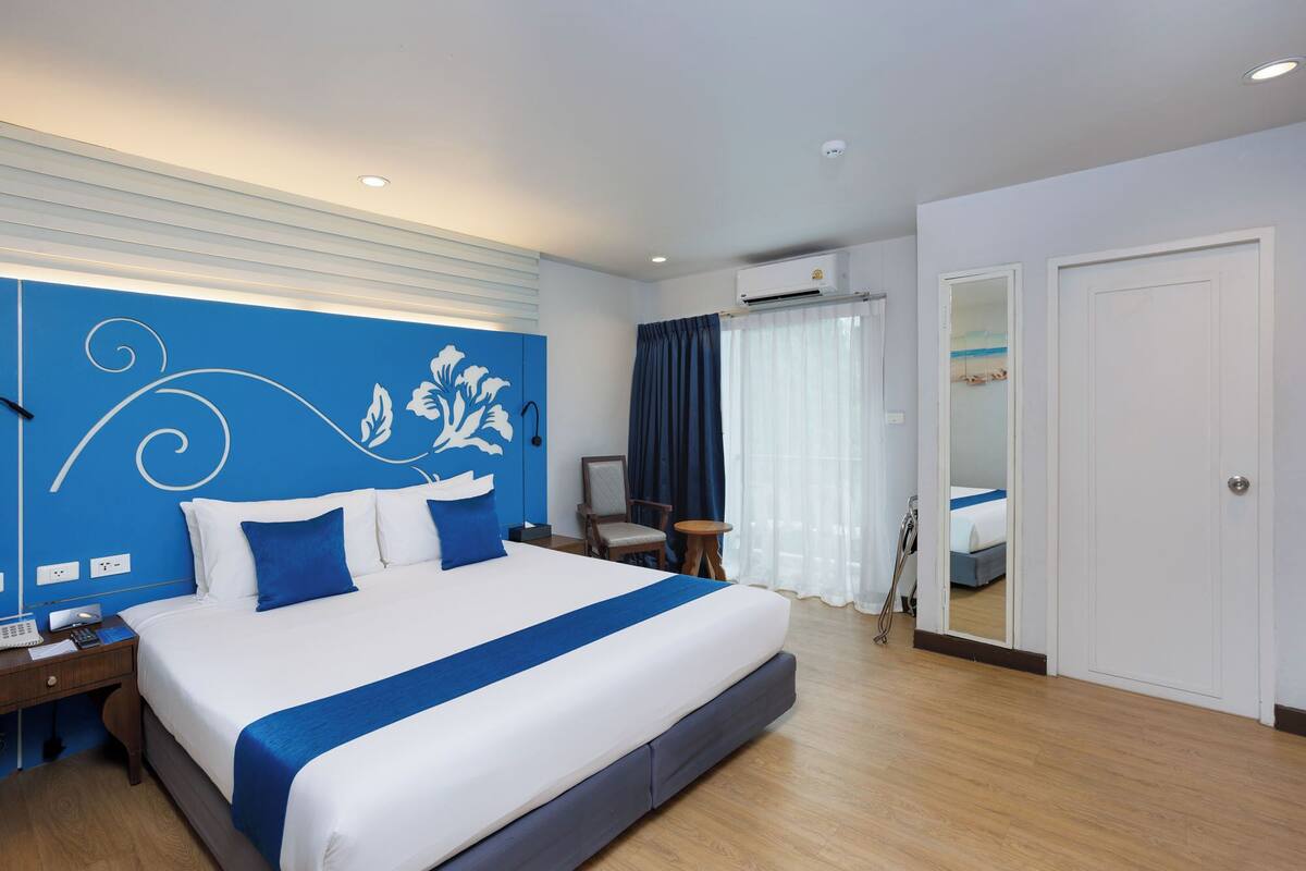 A hotel room with a large bed, a desk with a phone and papers, a chair, a small table, and a bathroom visible through an open door.