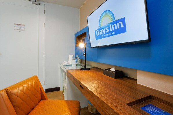 A hotel reception area with a television displaying the Days Inn logo, a wooden desk with a lamp, a cushioned orange chair, and a white door. - Days Inn by Wyndham Patong Beach Phuket (Patong)