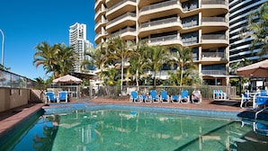 A swimming pool with blue chairs and umbrellas, surrounded by palm trees and a multi-story building with balconies. - Voyager Resort (Broadbeach)