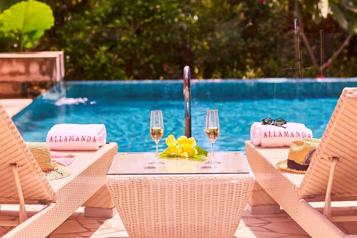 A poolside setting with two lounge chairs, a table with glasses of champagne, and towels with the name "ALLAMANDA".