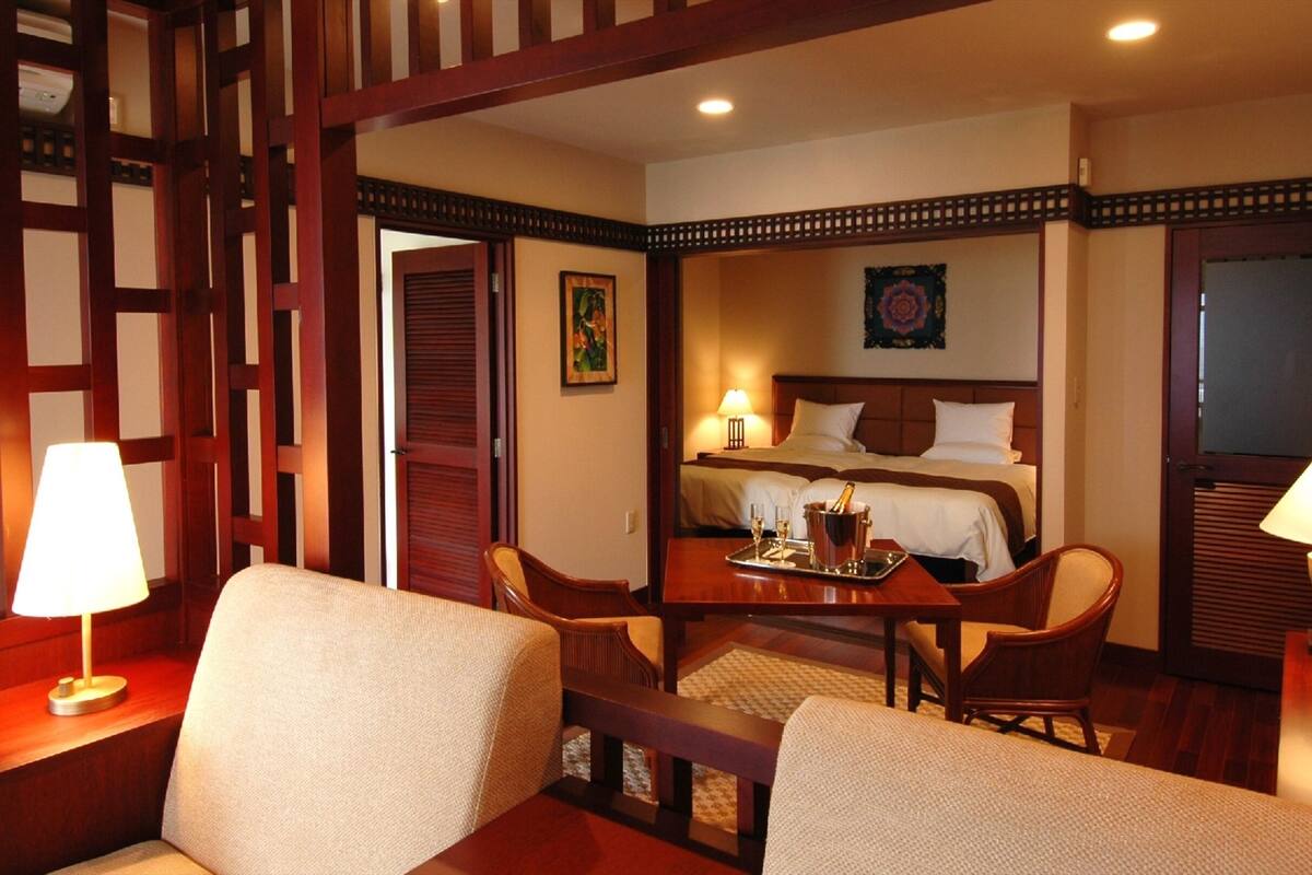 A hotel room with a bed, two chairs, a table with a tray of champagne glasses, and a lamp.