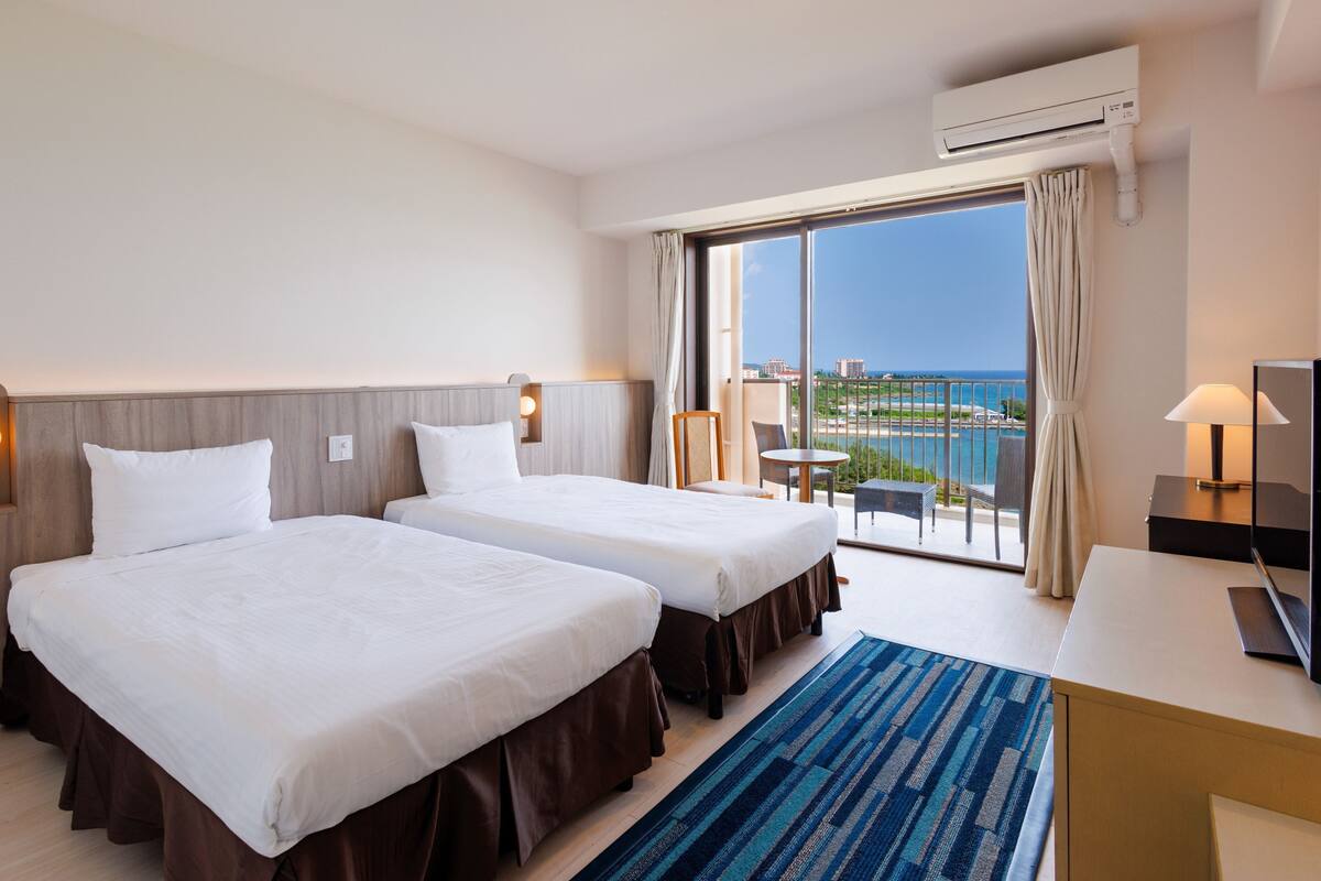 Ein Hotelzimmer mit zwei Betten, einem großen Fenster mit Meerblick, einem Balkon und einer Klimaanlage.