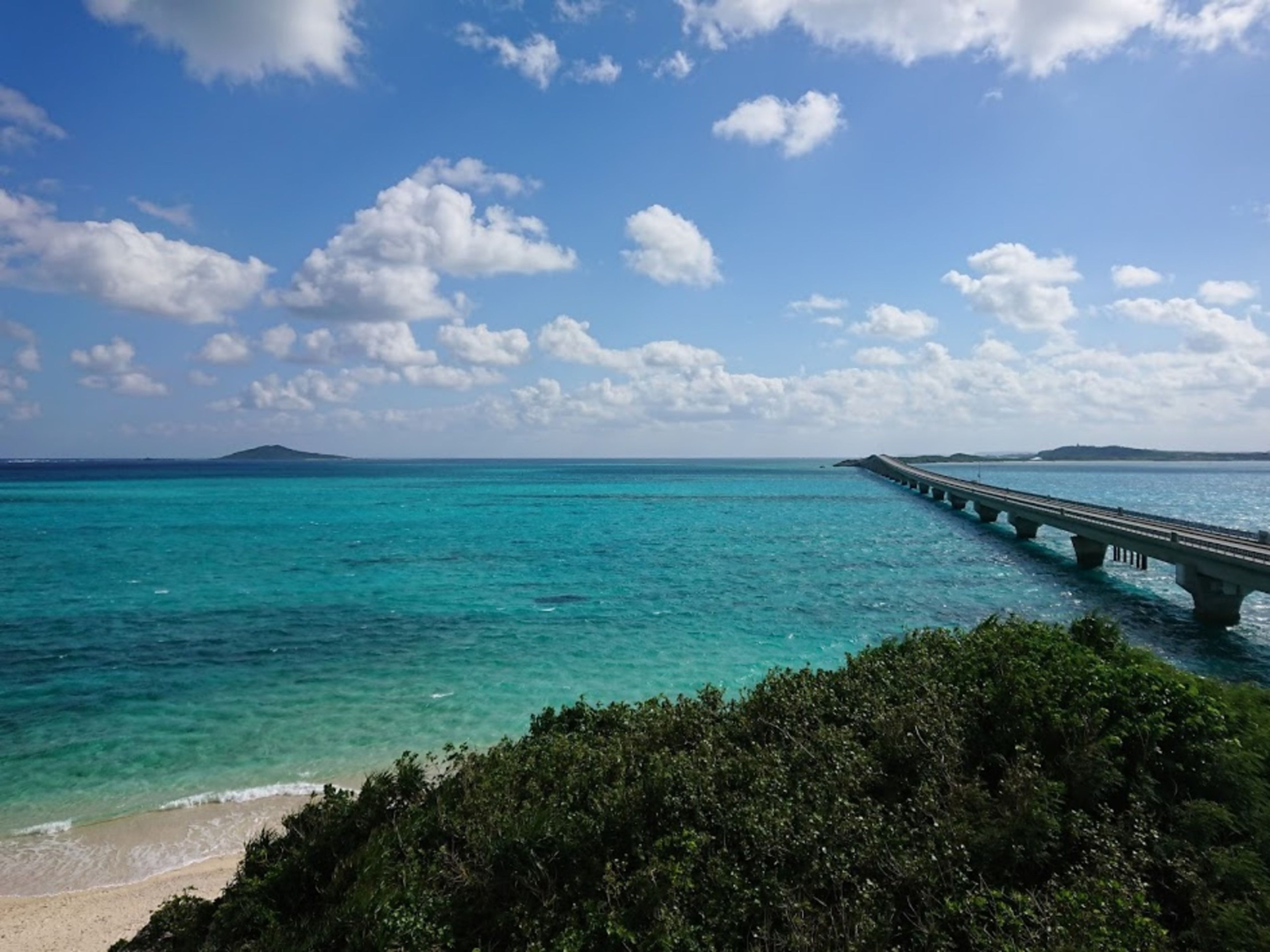 Escena costera con un puente que se extiende sobre aguas turquesas, una playa arenosa y abundante vegetación en primer plano.