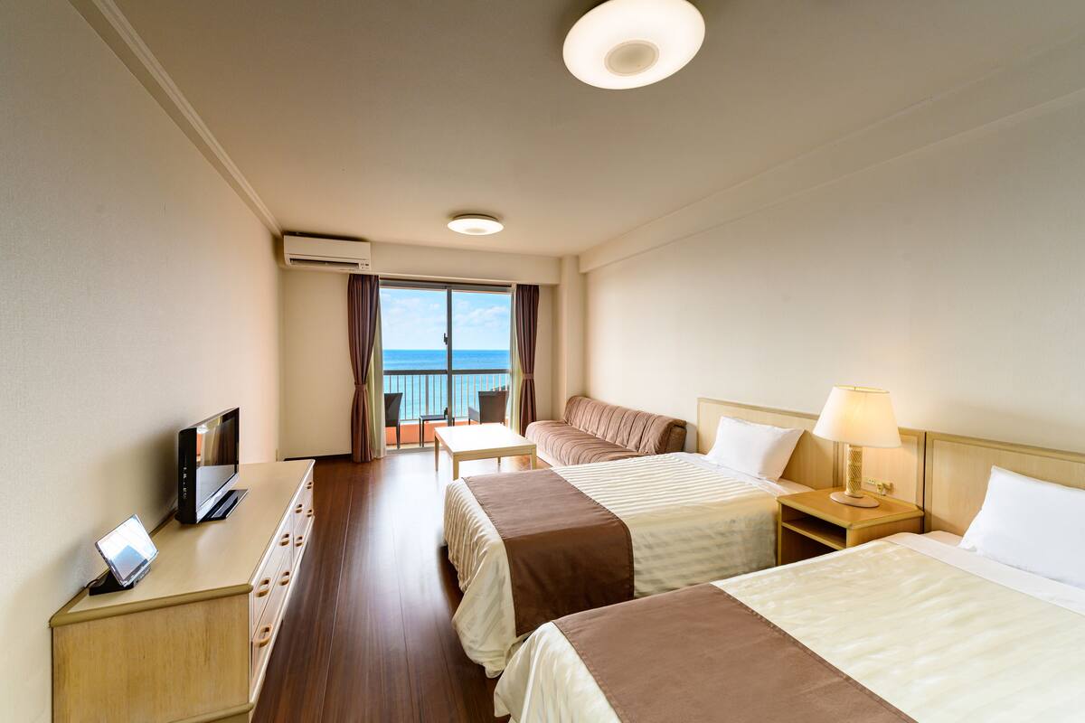 Ein Hotelzimmer mit zwei Betten, einem Schreibtisch mit Computer, einem Fernseher und einem Balkon mit Meerblick.