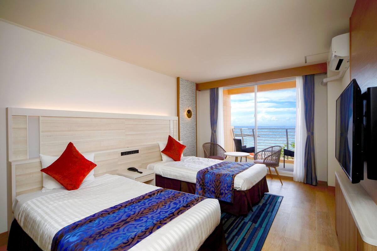 Ein Hotelzimmer mit zwei Betten, einem Fernseher, einem Balkon mit Meerblick und einem kleinen Tisch mit Stühlen.