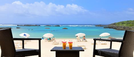 A beach view with two wicker chairs and a table, two umbrellas, and a clear blue sky.
