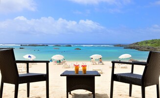 Vista de la playa con dos sillas de mimbre y una mesa, dos sombrillas y un cielo azul despejado.