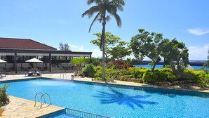 Una piscina con agua azul cristalina, rodeada de un patio con sombrillas y sillas, y vista al océano.