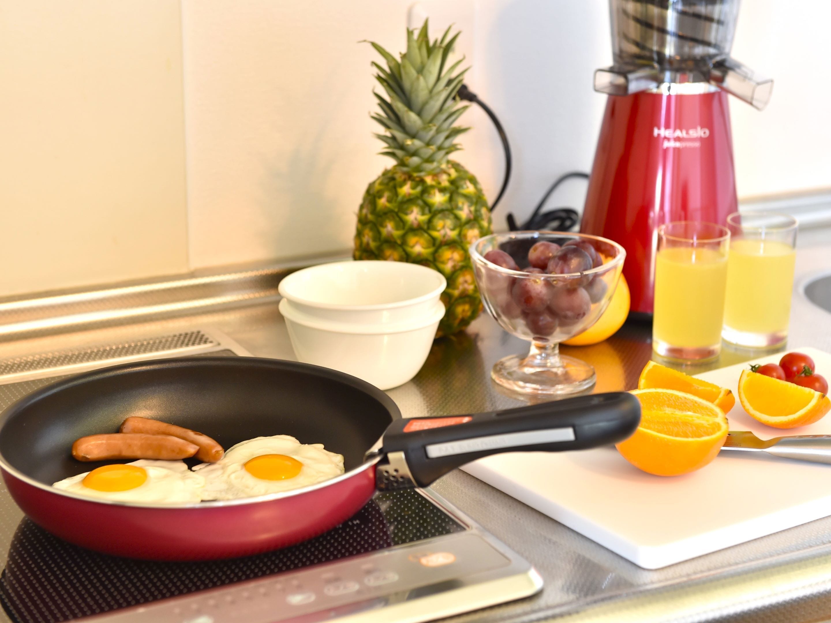Uma bancada de cozinha com uma frigideira contendo salsichas e ovos, uma abacaxi, uma tigela de uvas e copos de suco de laranja.