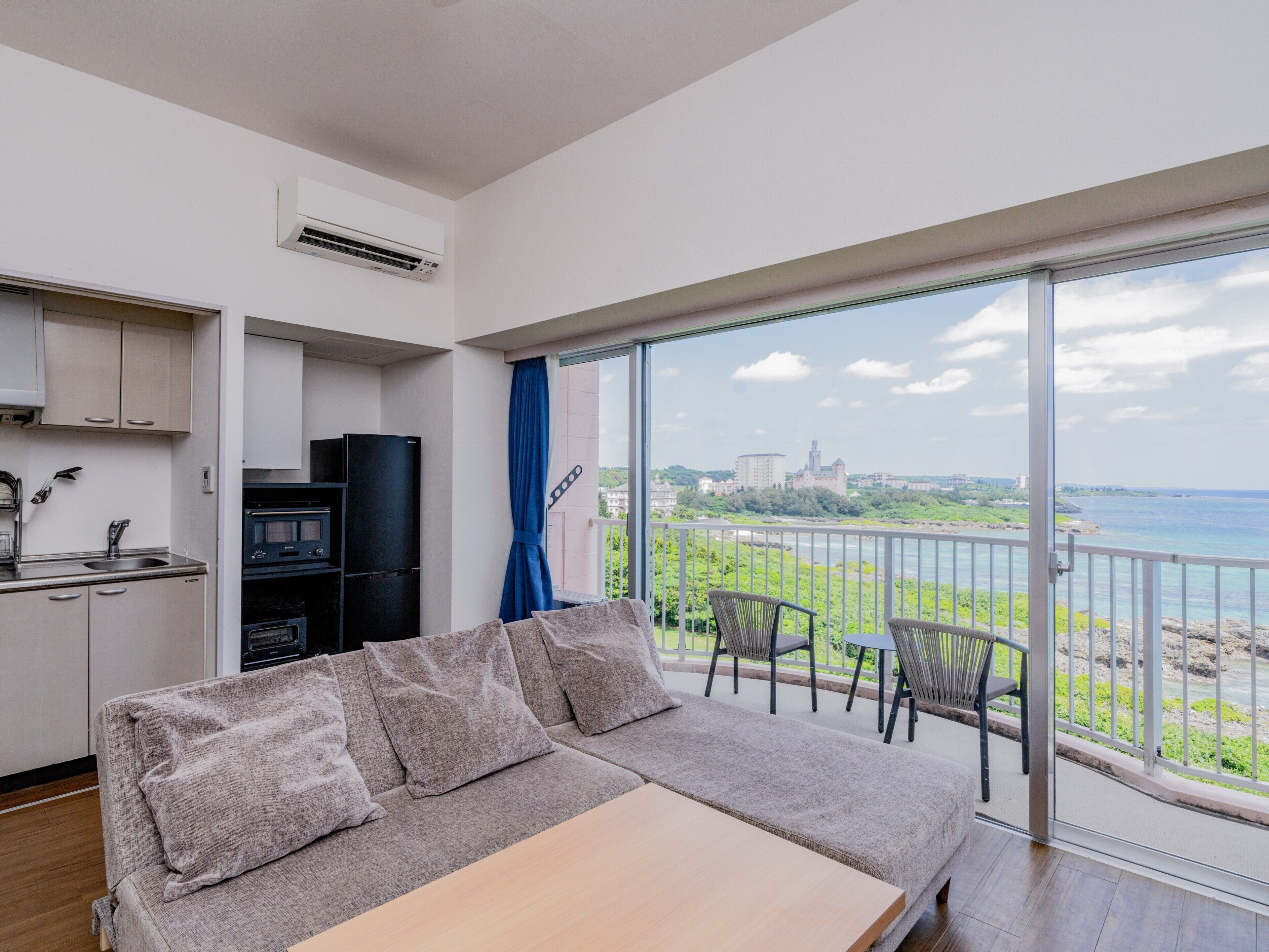A modern apartment with a kitchenette, a sofa, and a balcony with a view of the sea and a distant castle.