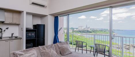 A modern apartment with a kitchenette, a sofa, and a balcony with a view of the sea and a distant castle.