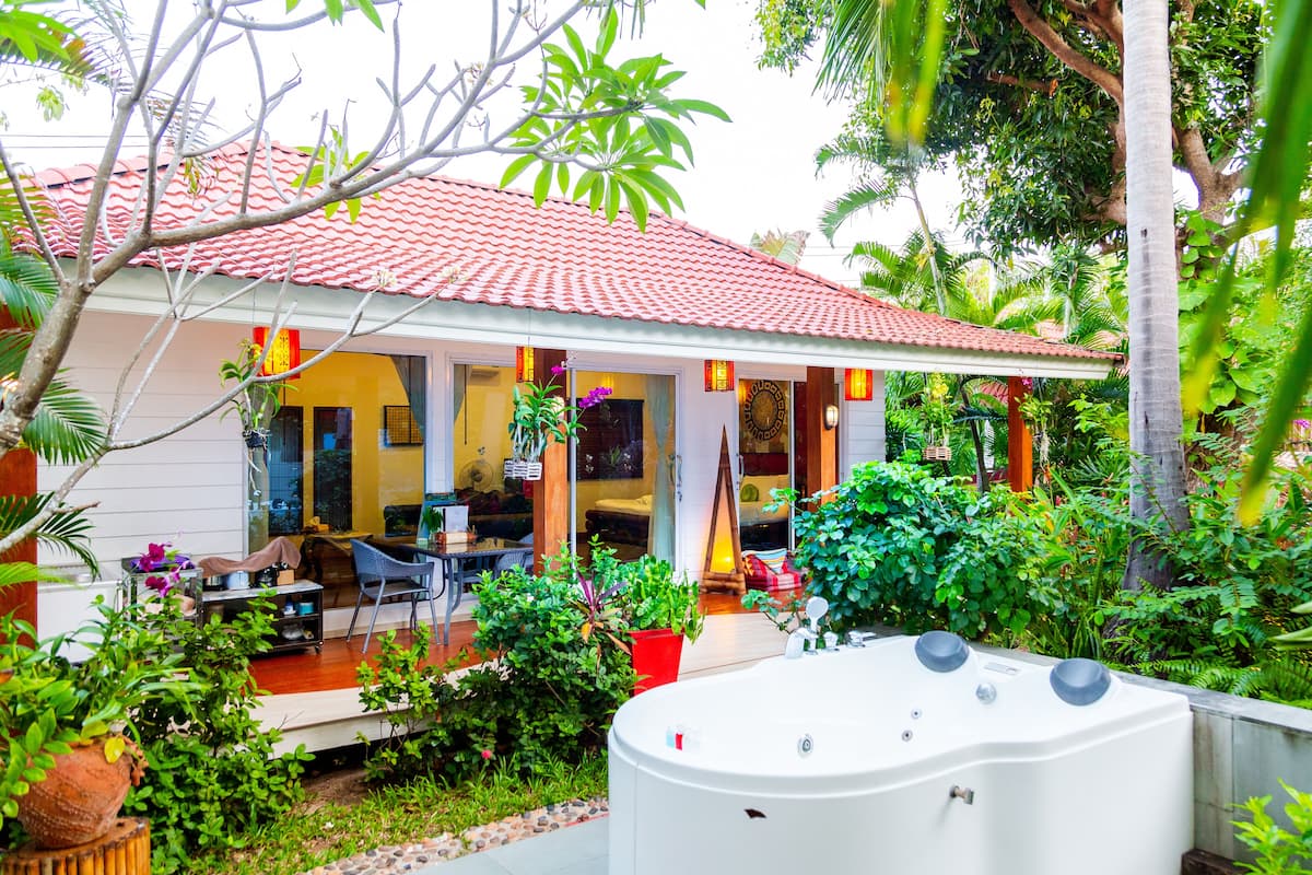 A house with a red tiled roof, a porch with chairs and a table, and a garden with various plants.