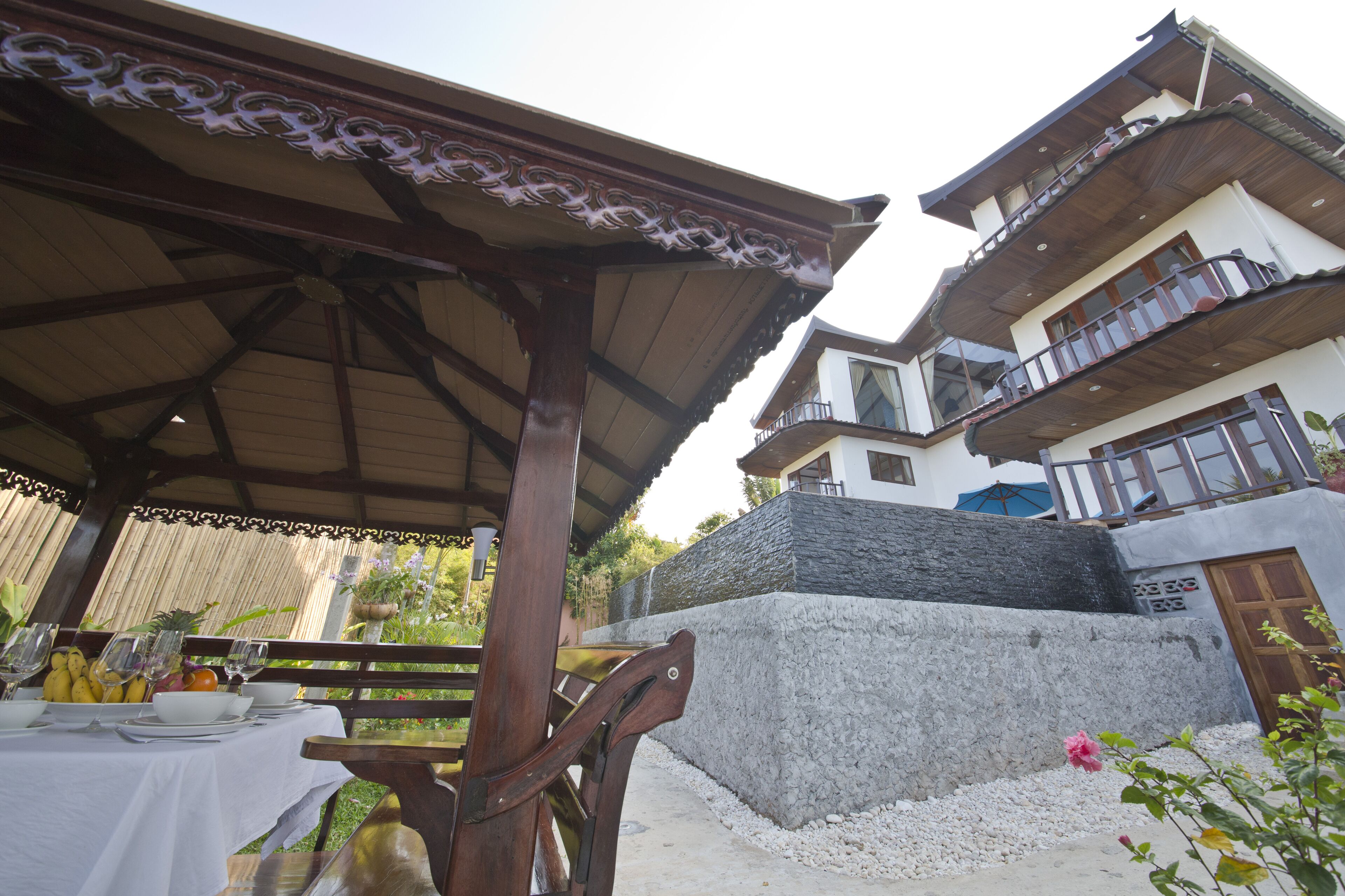 A covered outdoor dining area with a table set for a meal, a wooden bench, and a building with balconies and a stone wall in the background.