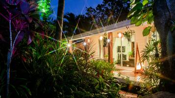 Una moderna abitazione con ingresso tramite una porta in vetro, circondata da rigogliosa vegetazione tropicale e illuminata da una calda luce interna.