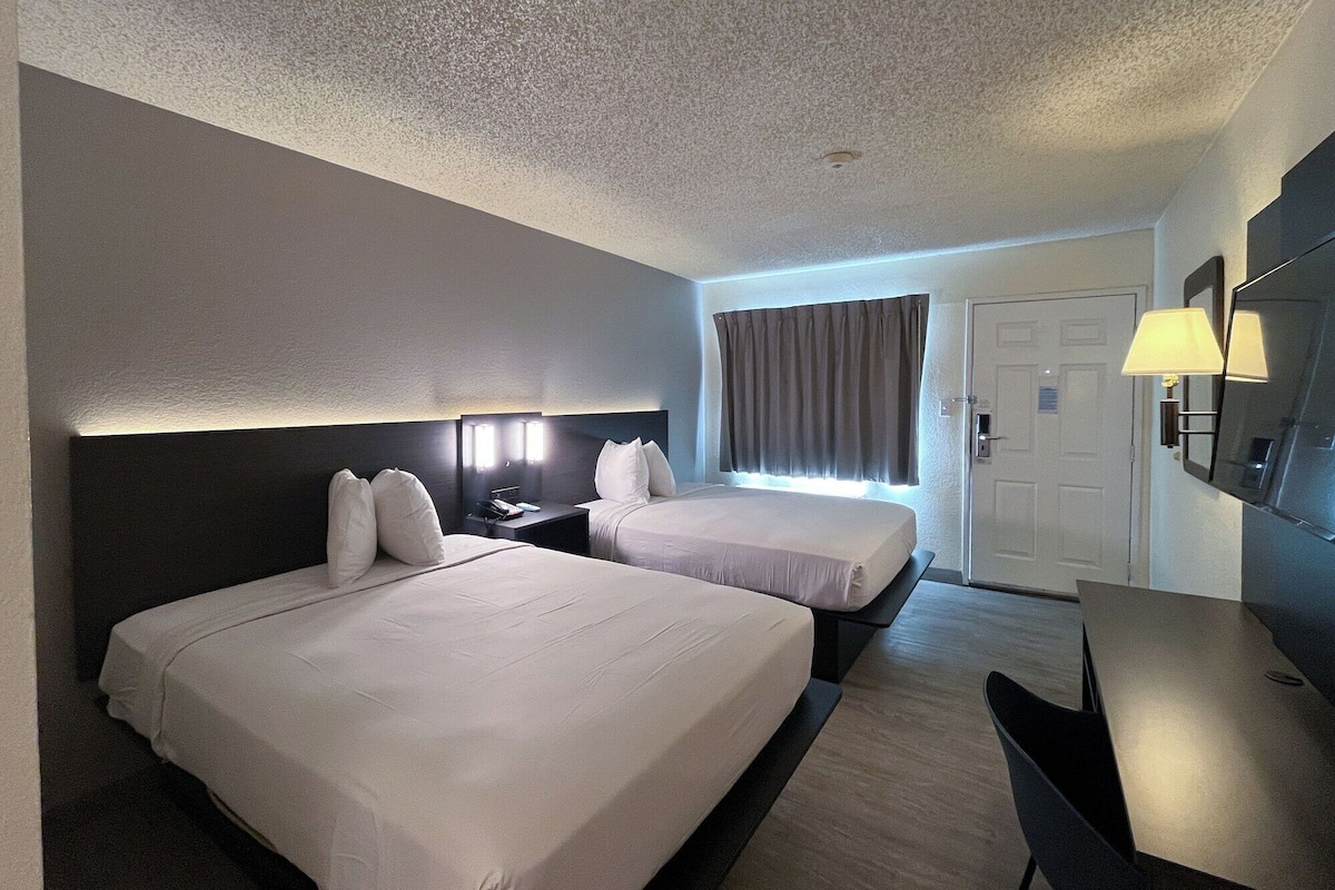 A hotel room with two beds, a desk, a chair, a TV, and a door.