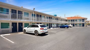 Un motel de varios pisos con una hilera de habitaciones en la planta baja, un estacionamiento y un cielo azul despejado.