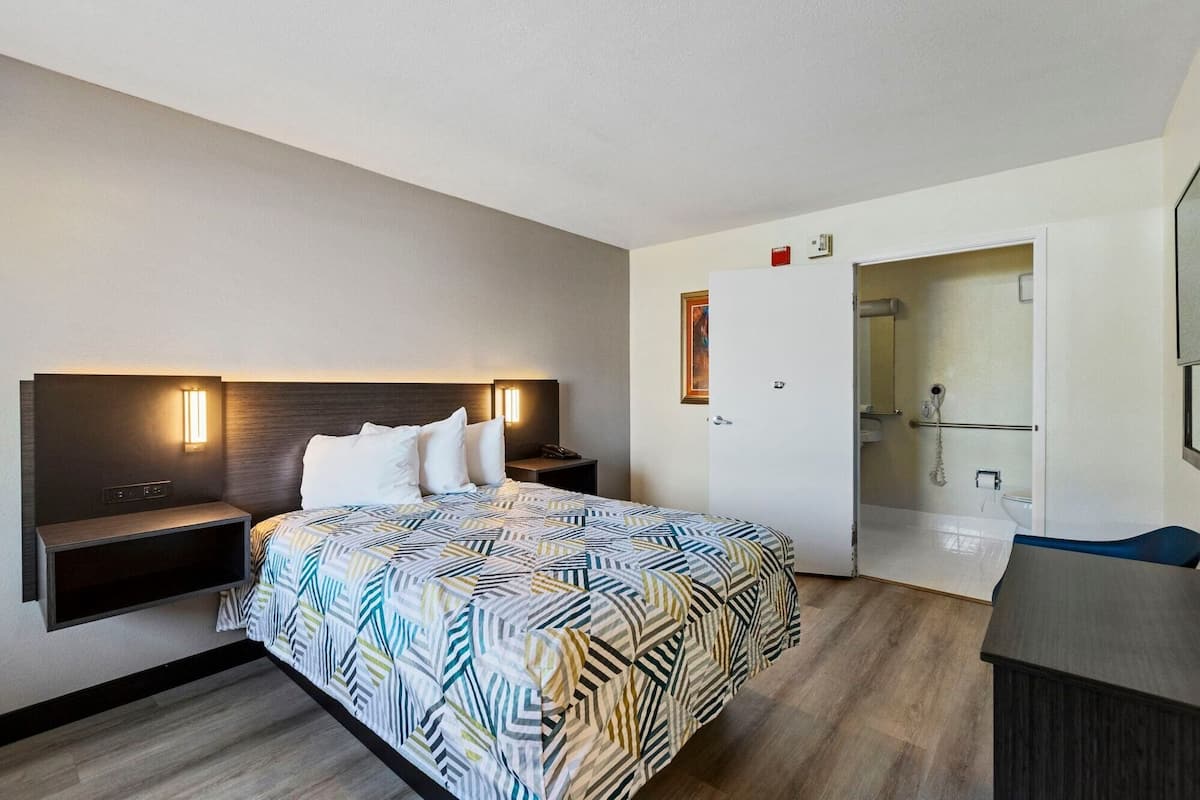 A hotel room with a bed, bedside tables, a wall-mounted TV, a door to a bathroom, and a shower area.