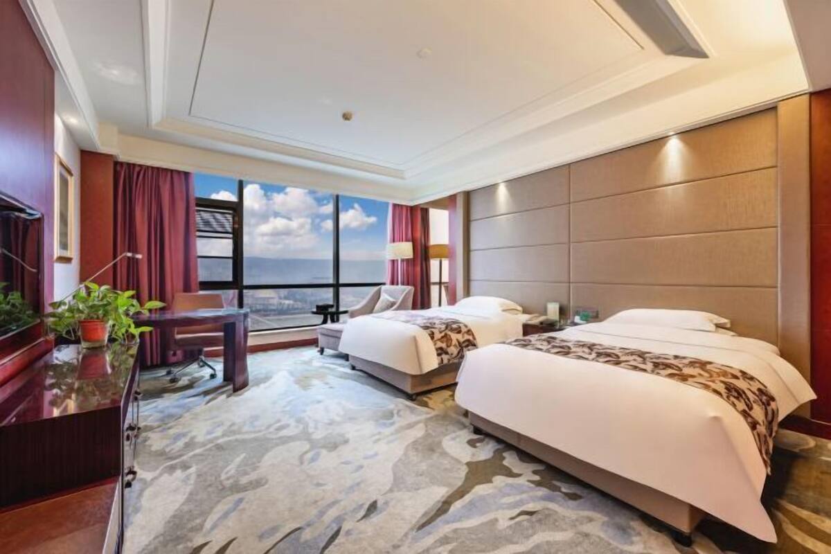 A hotel room with two beds, a desk, a chair, a TV, and a large window with a view.