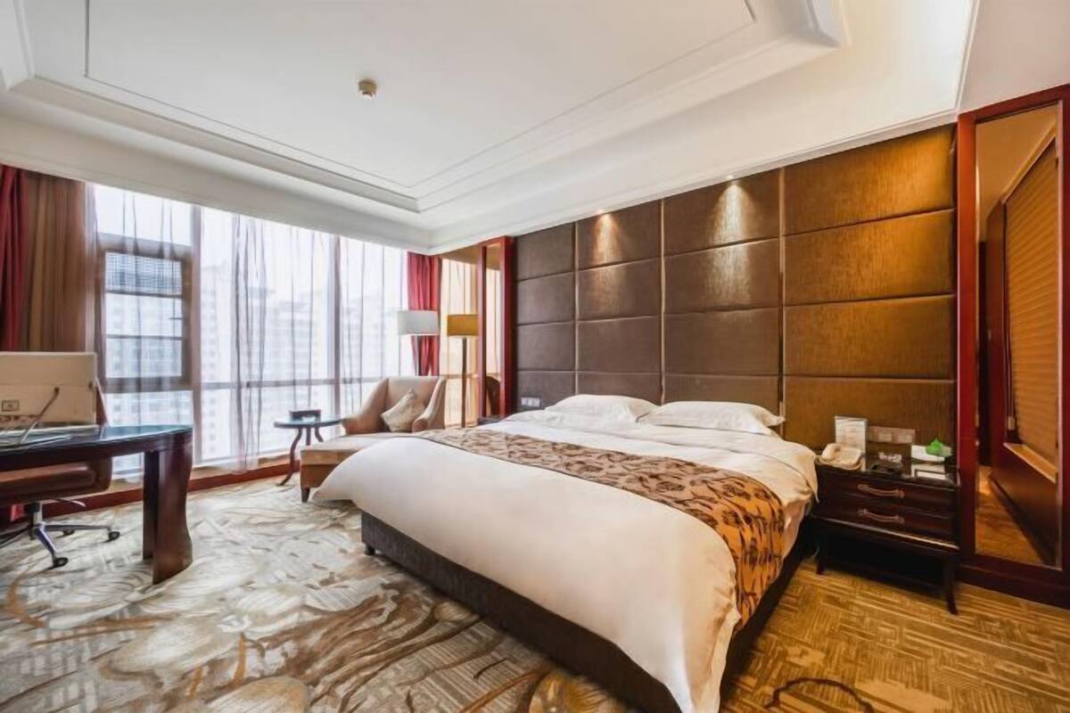 A hotel room with a large bed, a desk, a chair, and a view of the city through large windows.
