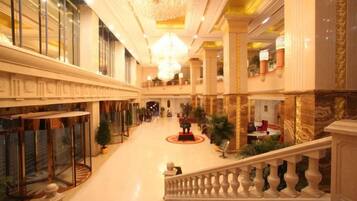 A grand hotel lobby with a central staircase, large chandeliers, and a spacious layout.