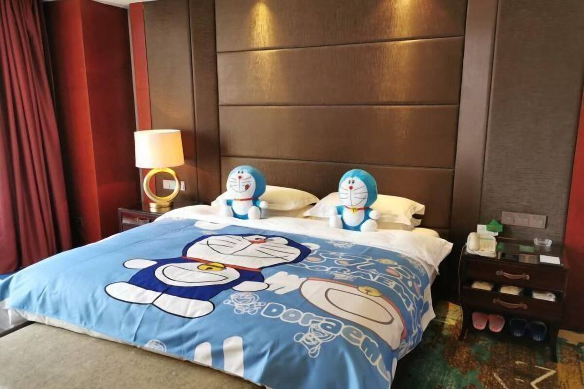 A hotel room with a large bed featuring a Doraemon-themed duvet, two plush toys on the headboard, a bedside lamp, and a nightstand with a lamp and a small drawer.