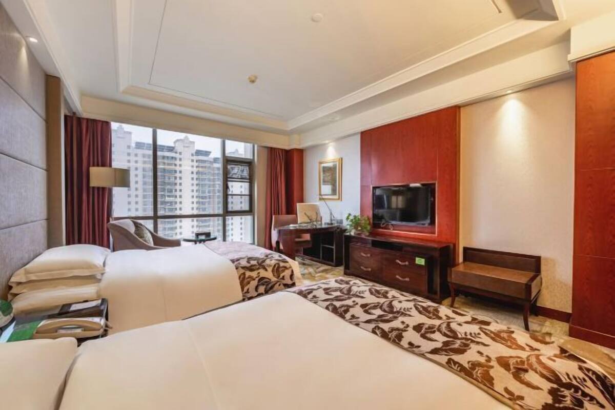 A hotel room with two beds, a desk with a TV, a window with a city view, and a red accent wall.
