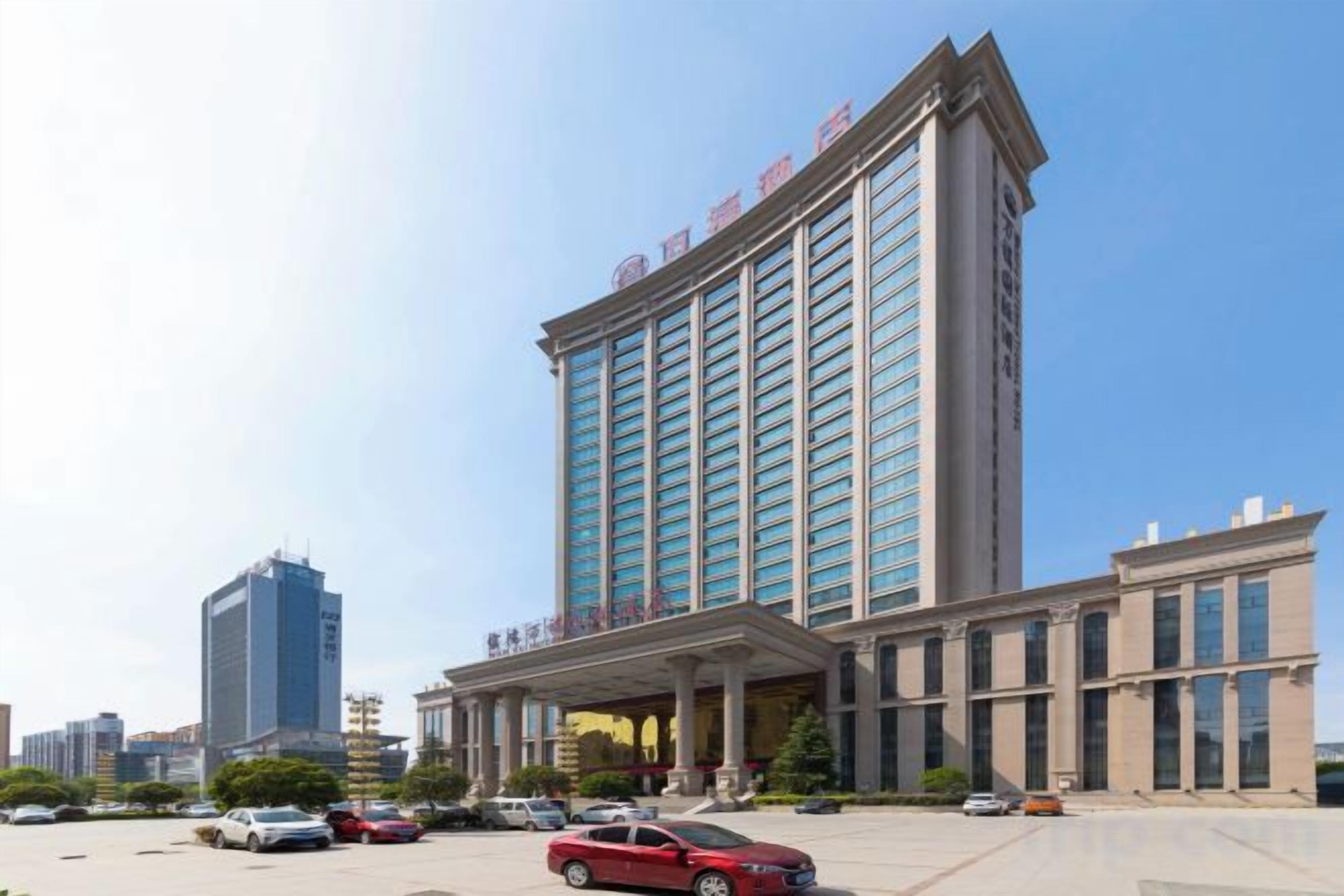 A multi-story hotel with a prominent entrance, large windows, and a parking area with several cars.